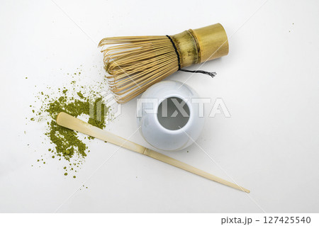 Traditional bamboo brush whisk with a spoon and matcha green tea powder splattered on white background. Accessories for making matcha green tea. Top view 127425540