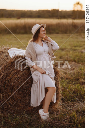 A woman stands in a summer field at golden hour leaning on a bale of hay. 127426362