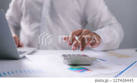 A businessman calculates financial data using a calculator and laptop on a desk filled with charts and reports. Ideal for illustrating accounting, budgeting, finance, and data analysis concepts. A businessman calculates financial data using a calculator and laptop on a desk filled with charts and reports. Ideal for illustrating accounting, budgeting, finance, and data analysis concepts. 127426445