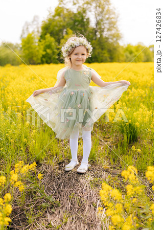Vertical portrait of cute preschooler girl wearing flower crown and elegant dress, joyfully dancing in vibrant meadow filled with bright yellow flowers on sunny summer day. Concept of happy childhood. 127426834