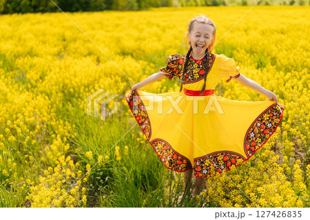 Cheerful young preteen girl in traditional Russian dress posing and smiling in vibrant field of yellow canola flowers on sunny summer day, embracing joy and beauty of childhood in nature landscape. 127426835