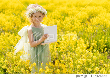 Charming little girl wearing fairy costume and flower crown holding blank canvas while standing in field of yellow flowers on sunny summer day, smiling looking at camera. Concept of happy childhood. 127426836