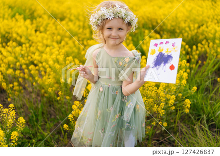 Portrait of cute talented child girl wearing flower crown and beautiful dress showing painting while holding brush standing in green meadow full of yellow flowers on sunny summer day, looking camera. Portrait of cute talented child girl wearing flower crown and beautiful dress showing painting while holding brush standing in green meadow full of yellow flowers on sunny summer day, looking camera. 127426837