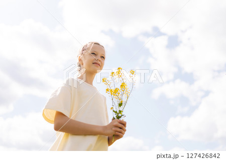 Shooting from below of pretty young girl with braided hair holding bouquet of yellow wildflowers standing posing in sunny summer meadow, enjoying beauty of nature under bright cloudy sky. 127426842