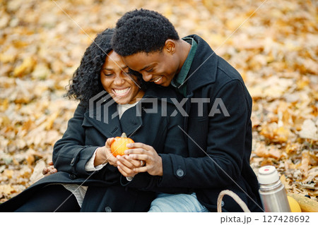 Black man and woman looking on little pumpkin. Romantic couple sitting on a blanket in autumn park. Man and woman wearing black coats. Black man and woman looking on little pumpkin. Romantic couple sitting on a blanket in autumn park. Man and woman wearing black coats. 127428692
