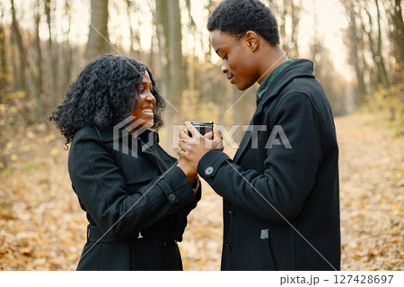 Black young man and his girlfriend holding in hands one coffee cup. Romantic couple walking in autumn park. Man and woman wearing black coats. 127428697