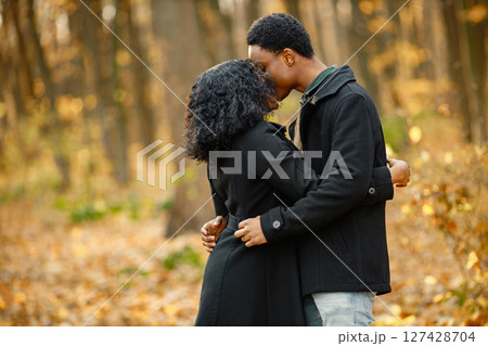 Black young man and his girlfriend kissing. Romantic couple walking in autumn park. Man and woman wearing black coats. Black young man and his girlfriend kissing. Romantic couple walking in autumn park. Man and woman wearing black coats. 127428704