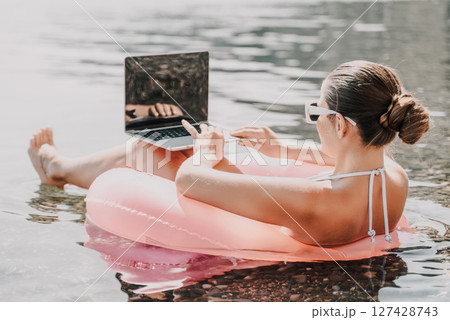 A woman is floating on a pink inflatable raft in the ocean while using a laptop. Concept of relaxation and leisure, as the woman enjoys her time in the water while working on her laptop. 127428743