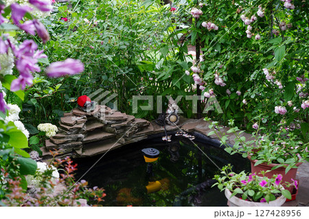 A lush flower garden surrounds a small decorative pond with stone edging, garden statues, blooming plants 127428956