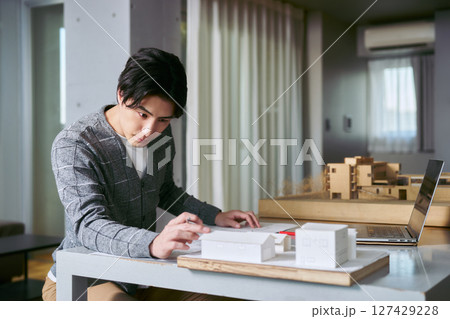建築模型を眺めながらノートパソコンと手元の図面で建物の意匠の設計の勉強をする建築学科の男性の学生 127429228