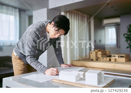 建築模型の置かれたデスクで設計図を確認し作図しながら設計を行う建築士の男性 建築模型の置かれたデスクで設計図を確認し作図しながら設計を行う建築士の男性 127429230