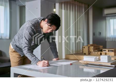 アトリエ系の設計事務所の建物の模型が置かれたデスクで立って図面を書く建築士の所員の男性 アトリエ系の設計事務所の建物の模型が置かれたデスクで立って図面を書く建築士の所員の男性 127429231