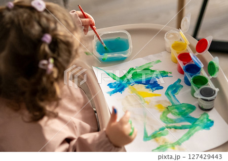 Top view of a young girl using a paintbrush with gouache while seated at a small art table.  127429443