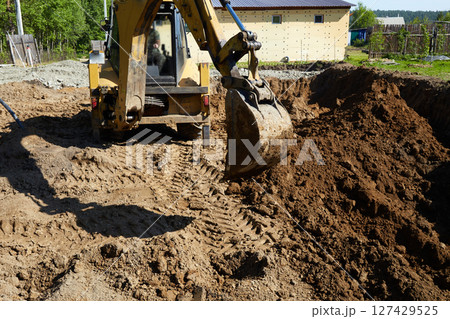 A digger operates in a rural setting, carefully digging a deep excavation for the construction of a house. A digger operates in a rural setting, carefully digging a deep excavation for the construction of a house. 127429525