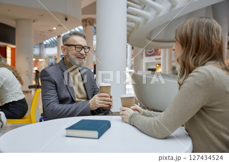 Mature man and young woman enjoying coffee and lively chats in a shopping centre Mature man and young woman enjoying coffee and lively chats in a shopping centre 127434524