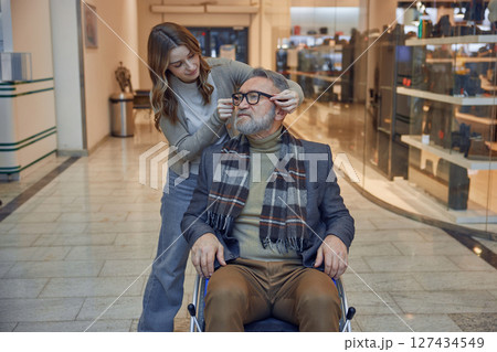 Woman assistant helping a mature man who uses a wheelchair in a shopping centre 127434549