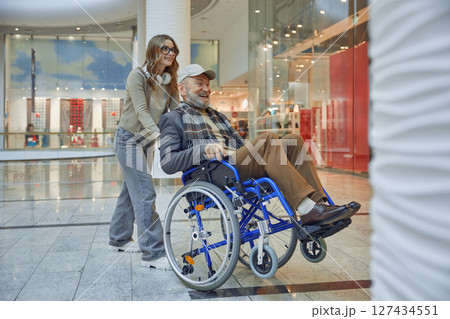 Granddaughter providing assistance to a senior man who uses a wheelchair at a shopping mall Granddaughter providing assistance to a senior man who uses a wheelchair at a shopping mall 127434551