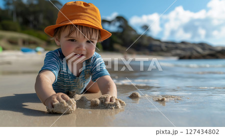 Smiling toddler boy in orange hat and striped shirt playing with wet sand at the beach near the water on a sunny summer day. Joyful childhood moment. 127434802