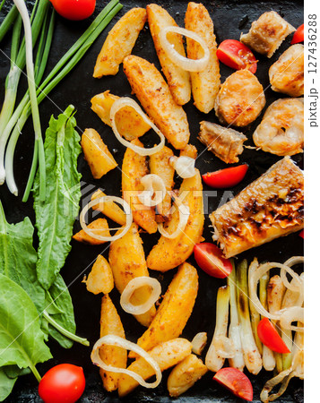 Oven pan with sliced baked potatoes, fish steaks and vegetables with onion on the table. Homemade healthy dinner with potatoes and seafood. 127436288