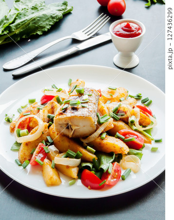 Plate with baked potatoes, piece of fish, vegetables and herbs on the table with tomato sauce. Dinner served with fish and chips meal. 127436299