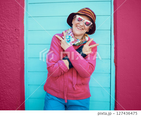 Portrait, peace and funky with a senior woman outdoor standing against a blue door and red wall background. Glasses, hands and hip hop with a happy mature female doing a hand sign or gesture outside 127436475
