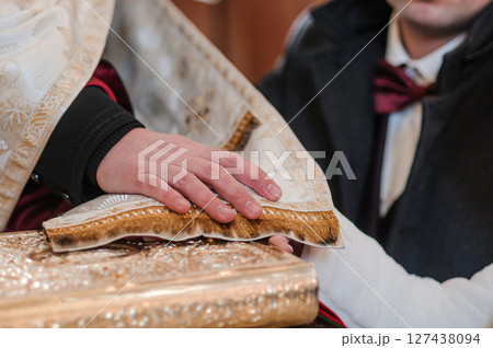 Solemn Eastern Orthodox wedding ceremony featuring a priest's hand holding a piece of bread over the hands of a groom dressed in a suit. Solemn Eastern Orthodox wedding ceremony featuring a priest's hand holding a piece of bread over the hands of a groom dressed in a suit. 127438094