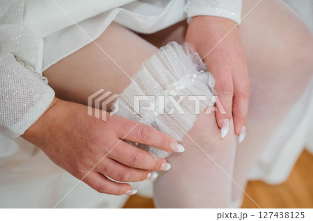 A close-up shot of a bride delicately holding a shimmering white lace garter in her hands, preparing for a wedding ceremony detail, with soft skin tones and delicate fabrics. 127438125