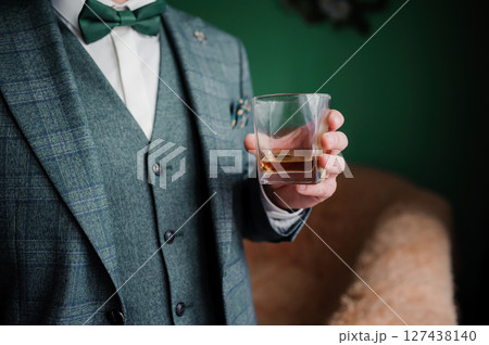 Stylish man in a green tweed suit with a bowtie holds a glass of whiskey, enjoying a drink in an elegant setting. Stylish man in a green tweed suit with a bowtie holds a glass of whiskey, enjoying a drink in an elegant setting. 127438140