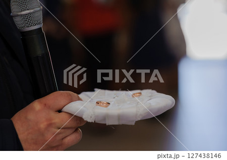 Wedding ring exchange ceremony focusing on a hand presenting a white cushion with two golden wedding rings, blurred guests. Wedding ring exchange ceremony focusing on a hand presenting a white cushion with two golden wedding rings, blurred guests. 127438146