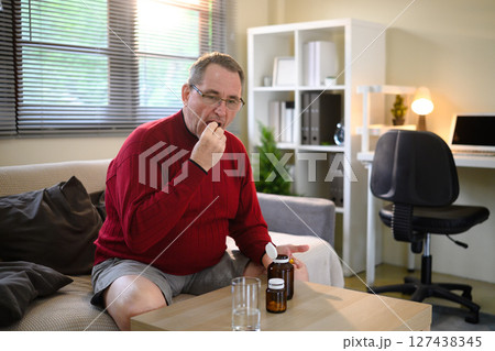 Senior man taking medication at home while sitting on the sofa in a cozy living room 127438345