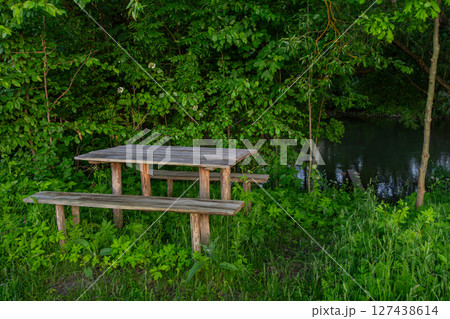Wooden picnic table and benches set in a serene green setting beside a calm stream during late afternoon Wooden picnic table and benches set in a serene green setting beside a calm stream during late afternoon 127438614