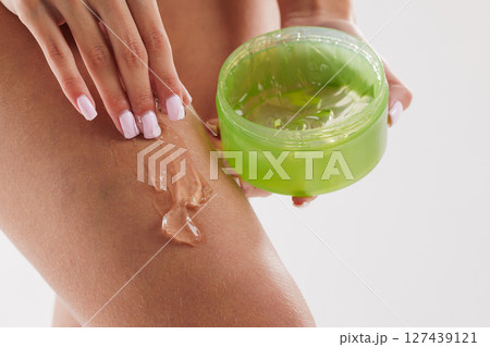 Close-up of woman applying clear gel from a green jar onto her leg, representing skincare, hydration, and body moisturizing routine on a white background. Close-up of woman applying clear gel from a green jar onto her leg, representing skincare, hydration, and body moisturizing routine on a white background. 127439121