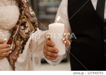 Romantic Wedding Candle Lighting Ceremony: Close-up of Hands Holding Lit White Pillar Candle, Bride's Lace Dress, Groom's Vest, Intimate Moment. 127440753