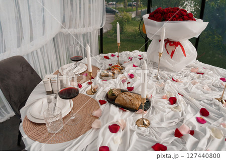 Romantic Dinner Date Setup: Table for Two with Red Roses, Wine Glasses, and Rose Petals in a Stylish Glass Room Setting Romantic Dinner Date Setup: Table for Two with Red Roses, Wine Glasses, and Rose Petals in a Stylish Glass Room Setting 127440800
