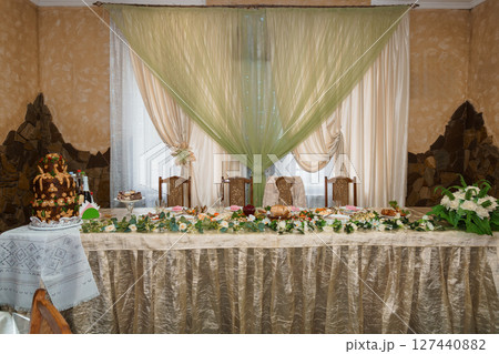 Luxurious Wedding Table Setting with Floral Decorations and Cake in Elegant Room Interior Featuring Beige Walls, Green Drapes, and Stone Accents. Luxurious Wedding Table Setting with Floral Decorations and Cake in Elegant Room Interior Featuring Beige Walls, Green Drapes, and Stone Accents. 127440882