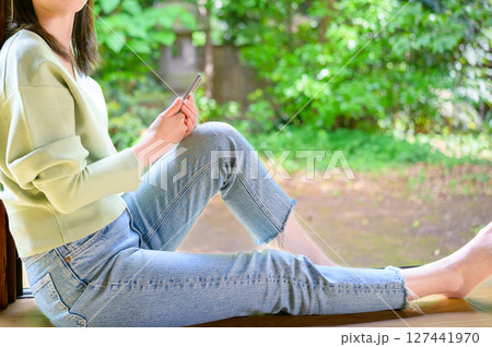 縁側で足を伸ばしスマートフォンを持つ女性が自然に包まれてくつろぐ夏の午後 127441970