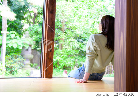 女性が縁側に寄りかかりながら木々を見上げる穏やかな夏の昼下がりのひととき 127442010