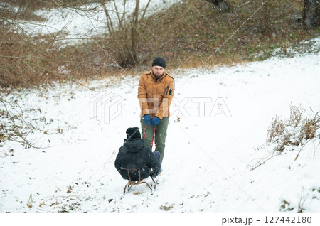A man pulls a child on a sled through a snowy landscape. Winter fun in the woods. A man pulls a child on a sled through a snowy landscape. Winter fun in the woods. 127442180
