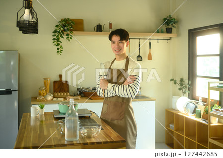 Confident young man wearing an apron and holding a spoon, standing in a home kitchen 127442685