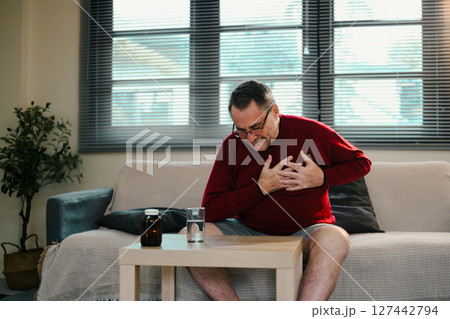 Senior man suffering from chest pain sitting on sofa at home, possibly indicating a heart attack Senior man suffering from chest pain sitting on sofa at home, possibly indicating a heart attack 127442794