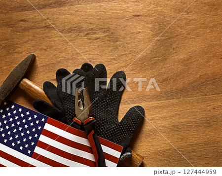 A textured wooden background with a pair of black work gloves, a hammer, and an American flag. A textured wooden background with a pair of black work gloves, a hammer, and an American flag. 127444959