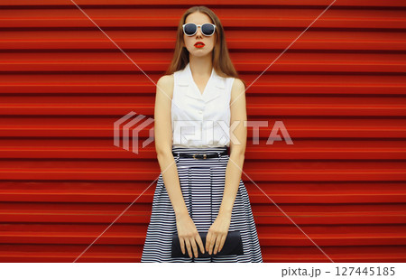 Beautiful stylish young woman with black handbag clutch, fashionable girl standing on red wall Beautiful stylish young woman with black handbag clutch, fashionable girl standing on red wall 127445185