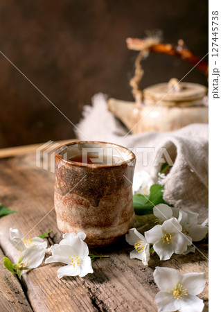 Ceramic tea cup with hot tea white spring flowers teapot on wooden table. Traditional oriental tea ceremony setup with natural decoration. Ceramic tea cup with hot tea white spring flowers teapot on wooden table. Traditional oriental tea ceremony setup with natural decoration. 127445738