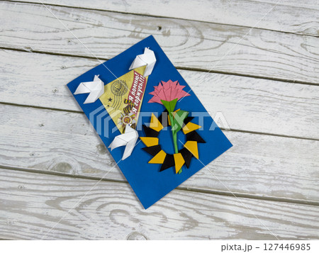 Victory Day greeting card with origami doves, pink carnation and yellow black laurel wreath on blue paper. Handcrafted tribute to WWII memory with rustic wooden background and vintage design Victory Day greeting card with origami doves, pink carnation and yellow black laurel wreath on blue paper. Handcrafted tribute to WWII memory with rustic wooden background and vintage design 127446985