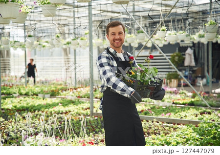 Black uniform, holding flowers. Man is working in garden center. Successful employee is in a bright greenhouse 127447079