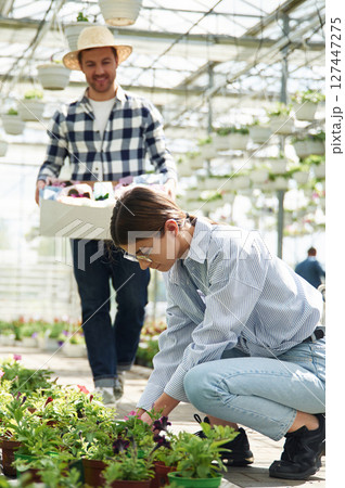 Man in straw hat is holding box with plants. Florist woman is working in bright greenhouse 127447275