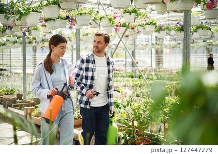 Walking, looking at each other, holding watering device. Florist man and woman are together in greenhouse 127447279