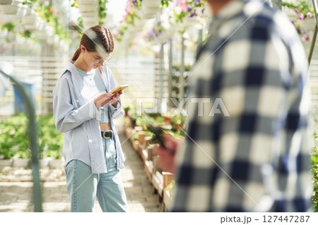 Holding smartphone. Florist man and woman are working together in bright greenhouse 127447287
