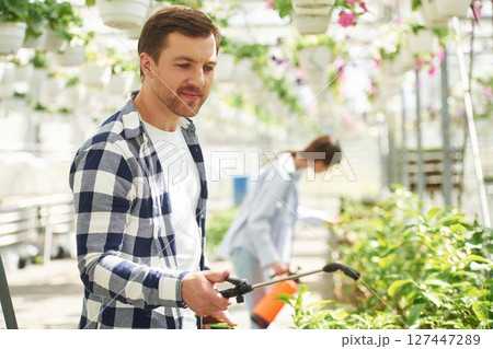 Holding watering cans. Florist man and woman are working together in bright greenhouse 127447289