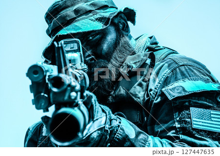 Soldier wearing camouflage and helmet aiming a rifle with intense focus, an American flag patch visible on uniform, set against a bright sky. 127447635